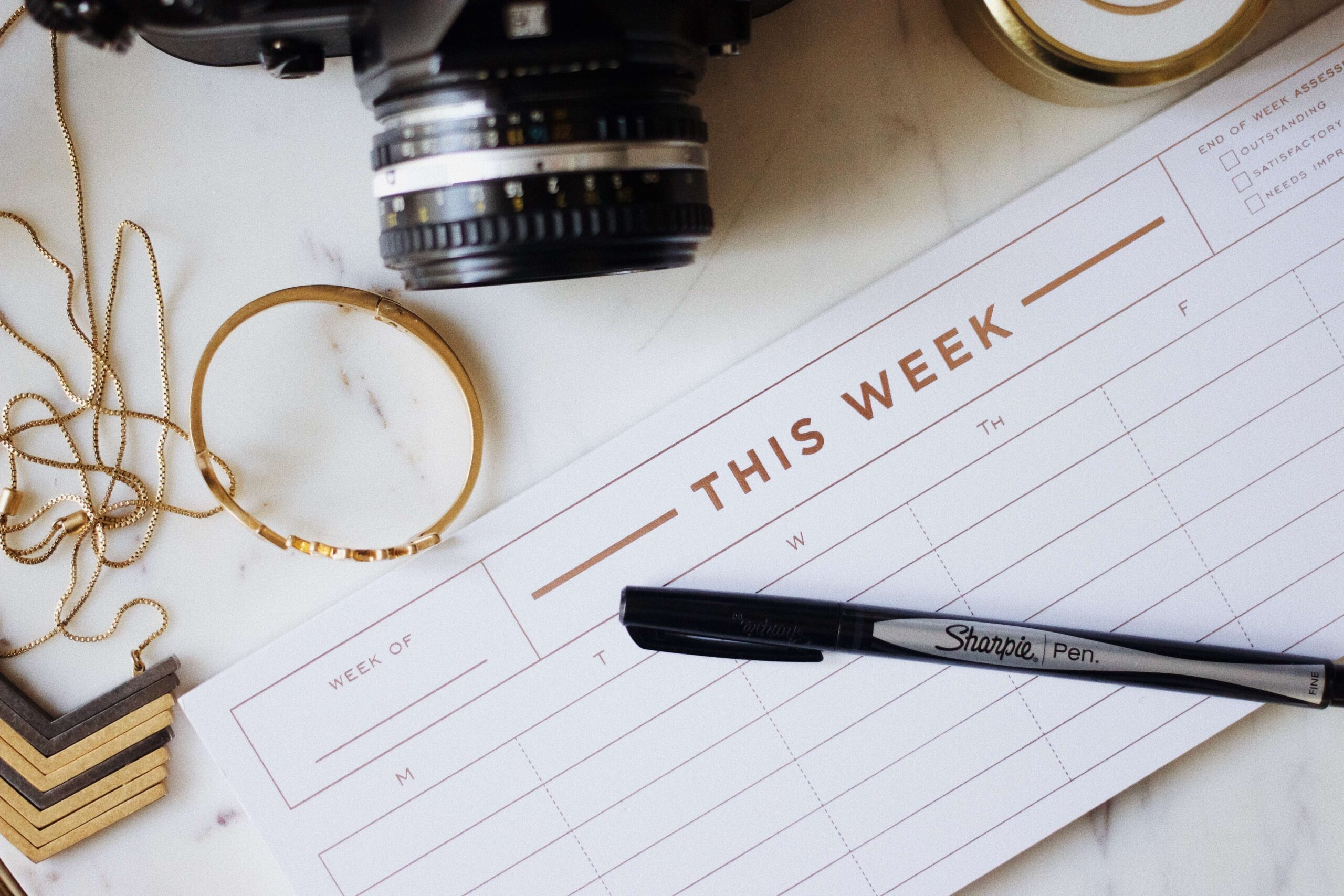 White pad planner with gold title, "This Week". A camera in the background. This post explains how to use a planner successfully